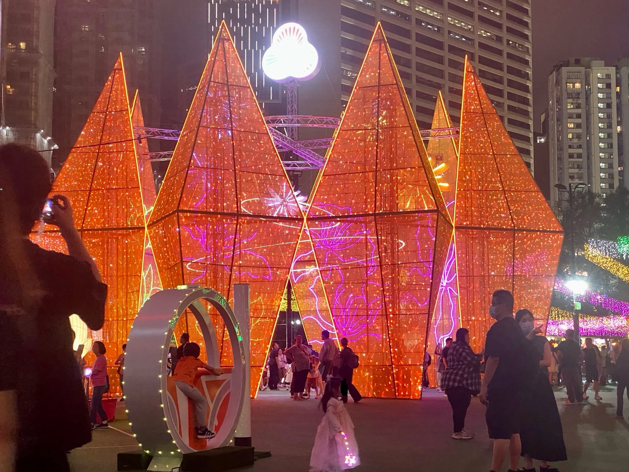 Large, sparkly, orange diamond-shaped displays adorned with pink and fuschia flowers.  Families in park.