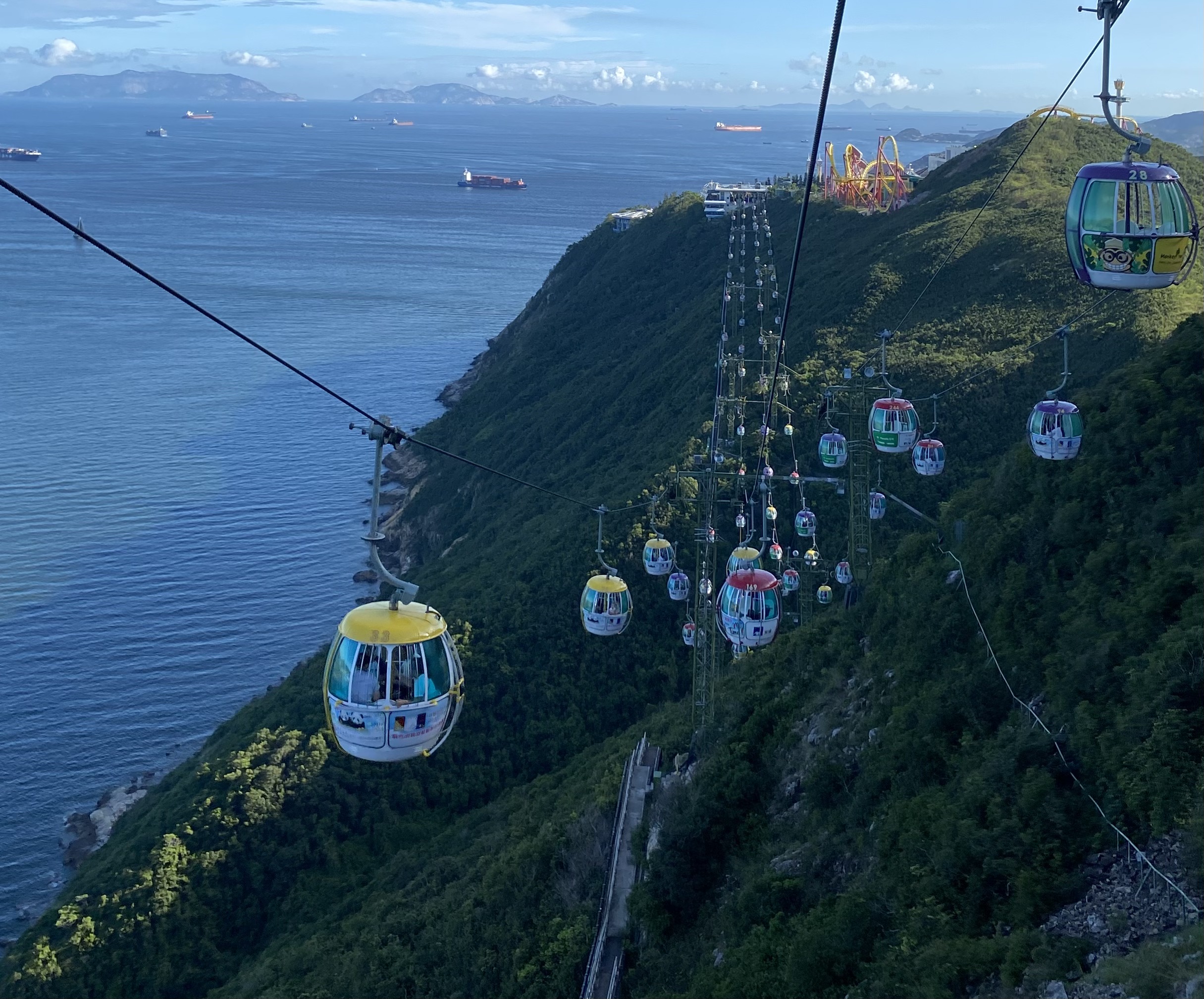 cable cars suspended in mid-air on the other side of the hill, a roller coaster in the distance, container ships in the sea as backdrop