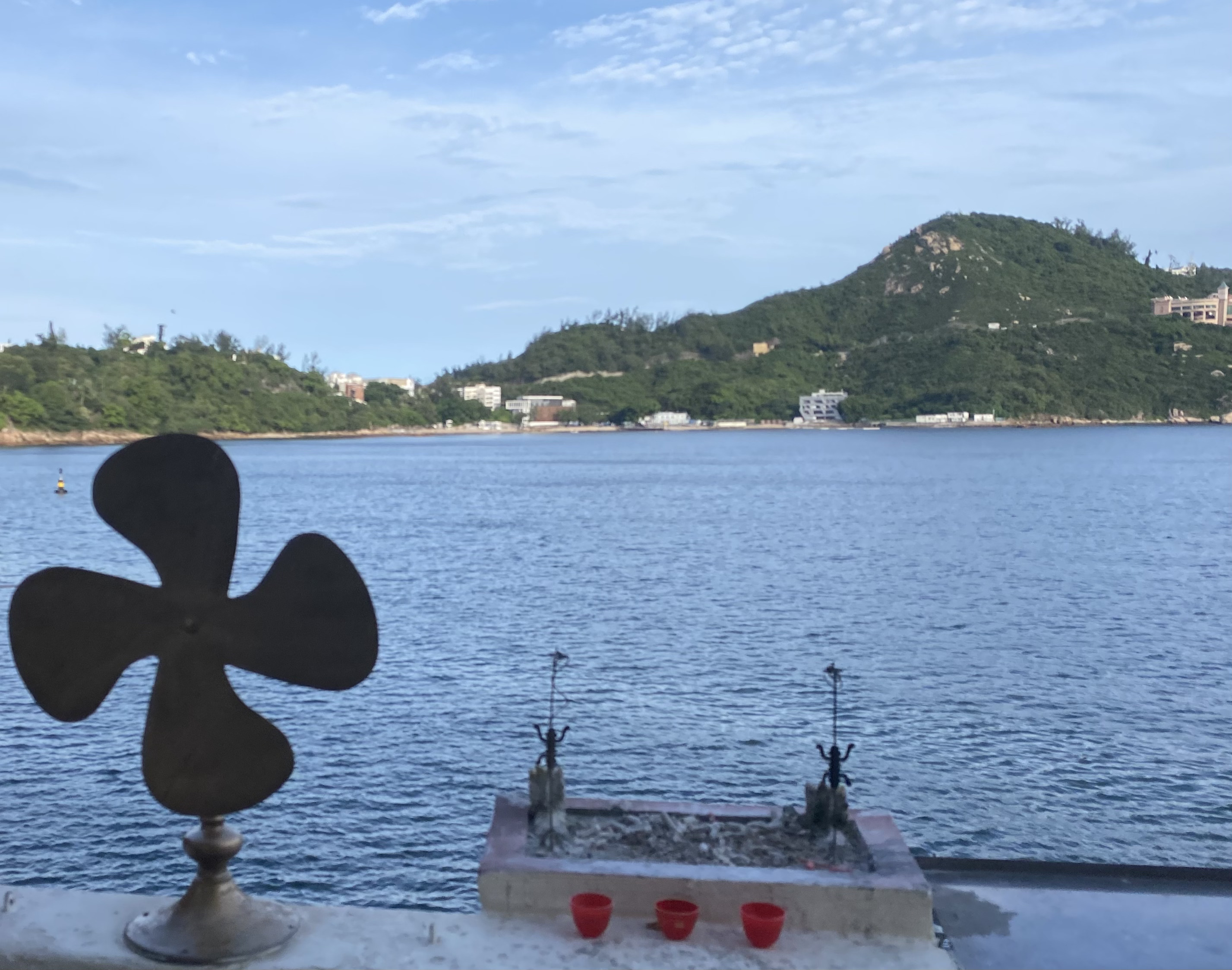 pinwheel and three tiny red cups in front of an incense holder on the waterfront with hills across the bay