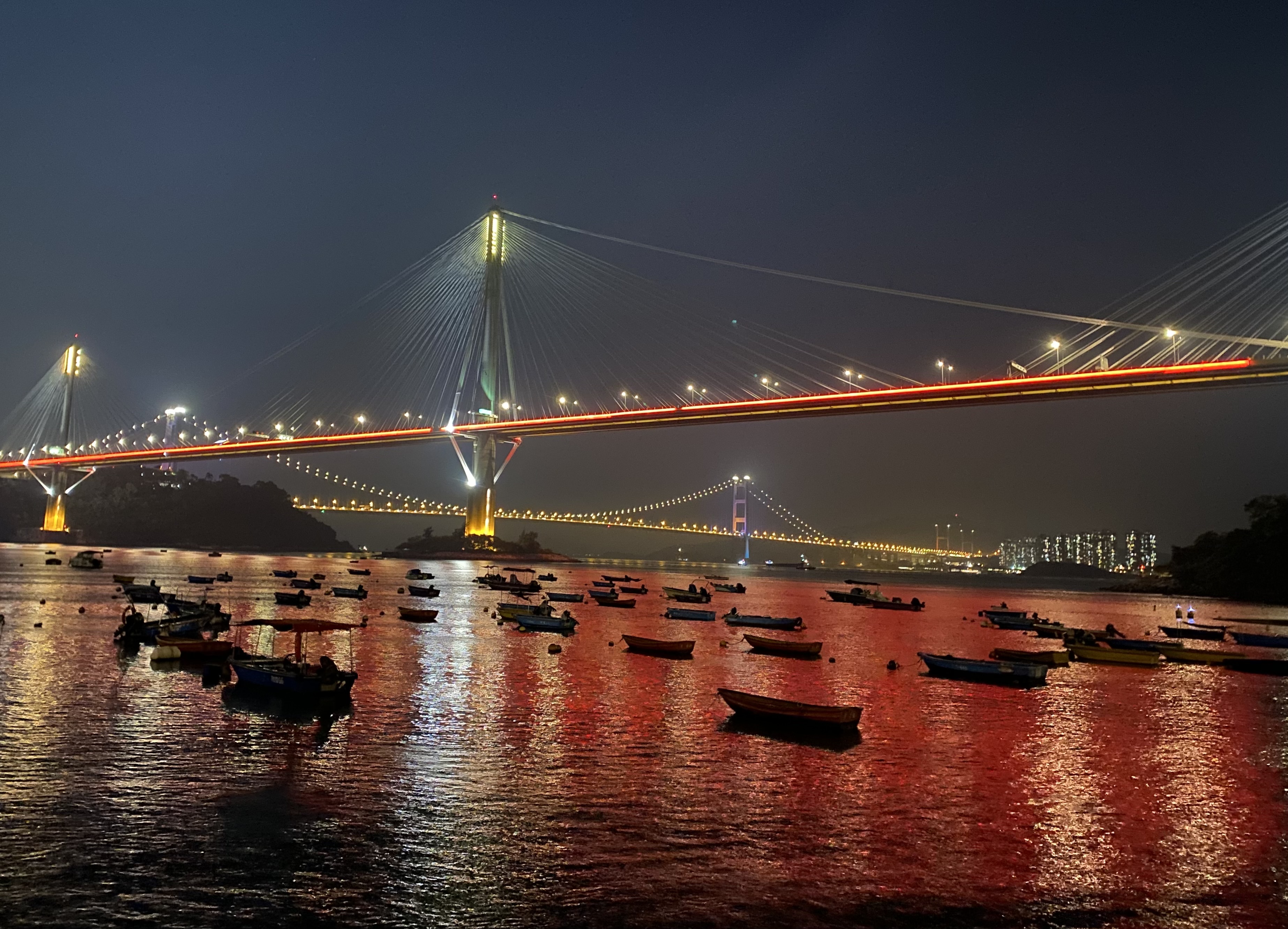 night scene of thirty or so small boats next to a bridge; another bridge in the distance